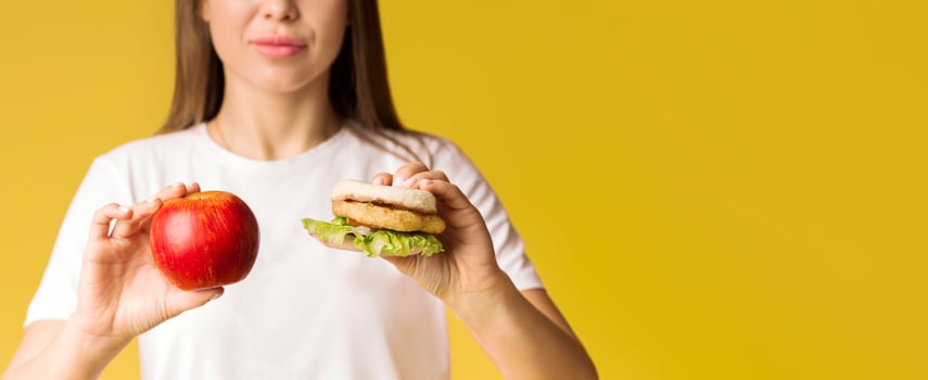 A woman stands confidently, holding an apple in one hand and a sandwich in the other, symbolizing the decision between healthy and indulgent food choices, copy space photo