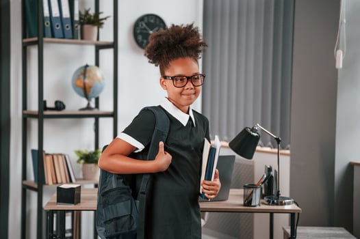 With backpack and notepad. Cute little girl in school uniform is standing indoors in domestic room photo