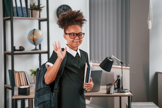 Bye, gesture. With backpack and notepad. Cute little girl in school uniform is standing indoors photo