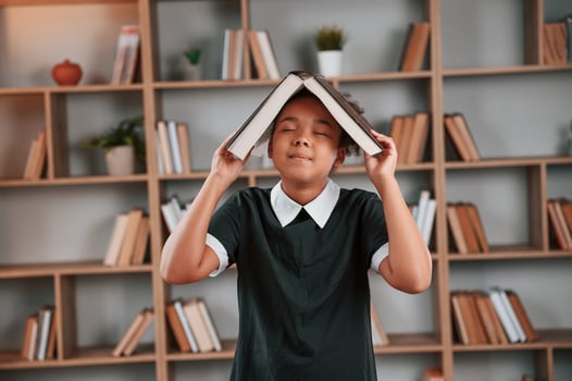 Having fun,, book is on the head. Cute african american girl in school uniform is at home library photo