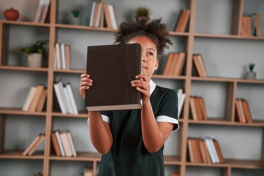Holding big book. Cute african american girl, in school uniform is at home library photo