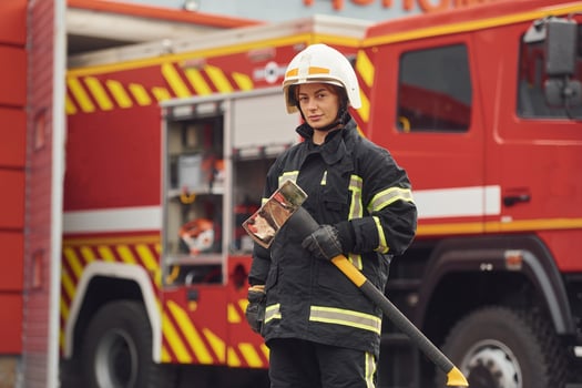 Standing, with axe in hands against truck. Woman firefighter in uniform is at work in department photo