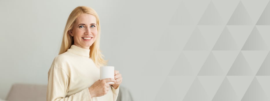 A young woman with long blonde hair enjoys a warm beverage while standing in a stylish indoor environment. Soft natural light fills the room, creating a cozy atmosphere, copy space photo