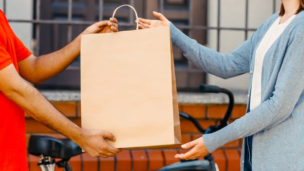 Food delivery, lunch to client and fast service. Millennial male courier sits on bicycle in company t-shirt, giving order to woman client in eco paper bag with order near house, cropped, panorama photo