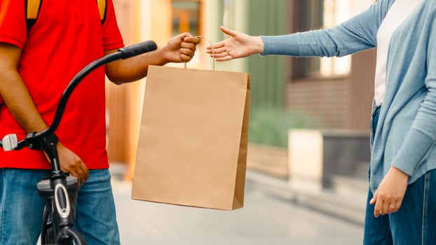 Male courier with bicycle delivered no name shopping bag to client and food service at home. Young guy in orange t-shirt giving paper eco bag to female near house, cropped, outdoor, empty space photo