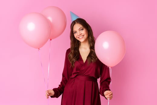 A cheerful young woman stands against a pink backdrop, holding three pink balloons while wearing a party hat. She radiates happiness, embodying the spirit of celebration. photo