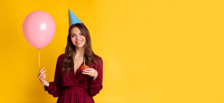 A young woman in a burgundy dress smiles while holding a pink balloon and a piece of cake. She wears a festive party hat, embodying the cheerful spirit of a birthday celebration, copy space photo