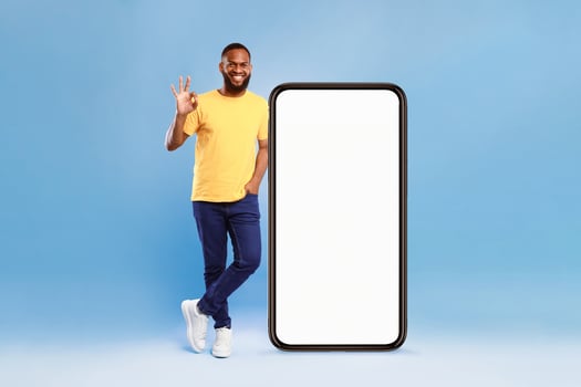A cheerful African American man, casually dressed in a yellow t-shirt, stands next to a large blank smartphone. He shows a hand sign, exuding positivity against a vibrant blue background. photo