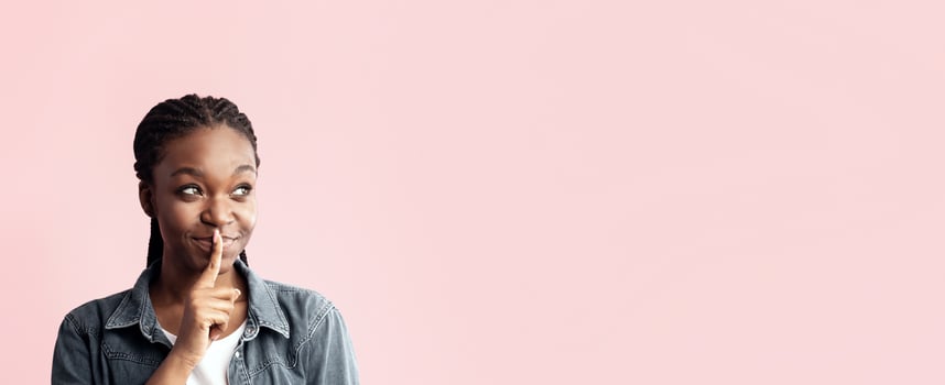 A young woman with braided hair playfully gestures for silence with her finger to her lips, conveying a sense of secrecy or fun. The soft pink background enhances her expression. photo