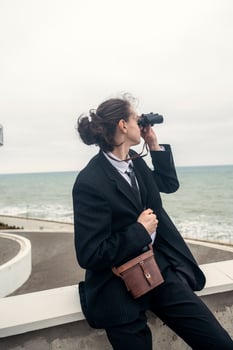 A young man sits alone against the backdrop of modern architecture near the sea. He looks through binoculars at the sea. He has long hair, wears a suit and tie. Relaxed, peaceful. free on the seashore photo