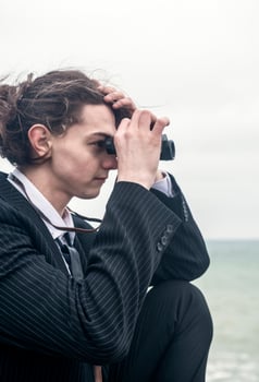 Portrait of a young man against the backdrop of the sea, he is looking into the distance through binoculars. He has long hair, is wearing a suit and tie. Relaxed, peaceful, thoughtful, happy and free on the seashore. photo