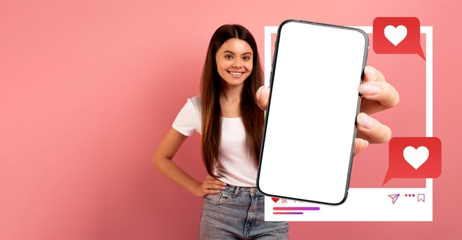 A woman stands against a pink background, smiling while holding up a smartphone. The device displays a blank screen, ideal for showcasing social media applications and engagement. photo