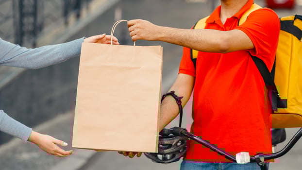 Goods delivery at city. Girl takes a paper bag from courier with big yellow backpack and bicycle outdoors, cropped, close up, free space photo