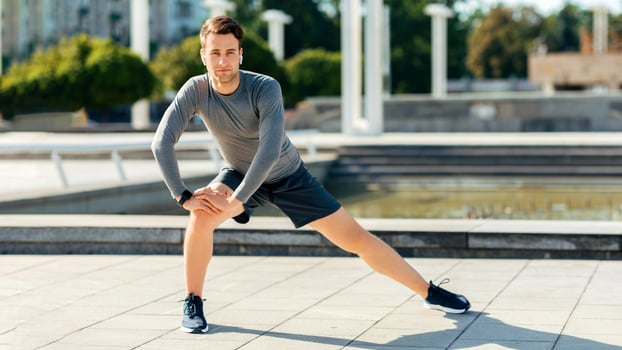 Training before marathon. Young man with wireless headphones in sport uniform doing exercises for legs, free space photo