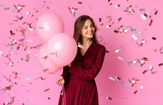 A young woman in a burgundy dress smiles while holding three pink balloons amid a flurry of colorful confetti. The vibrant pink background enhances the celebratory atmosphere. photo