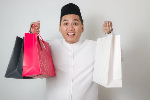 close up of cheerful happy young asian menl wearing modern ramadan outfit and eid mubarak idul fitri, holding shopping bags looking at camera photo