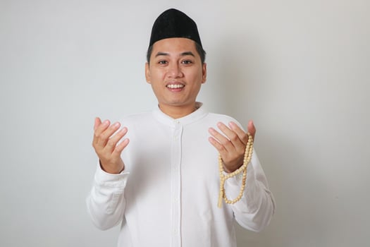 Portrait Of Young Asian Man Wearing Islamic Clothes Smiling While HOlding Prayer Beads Or Tasbeeh Isolated On Beige Background photo