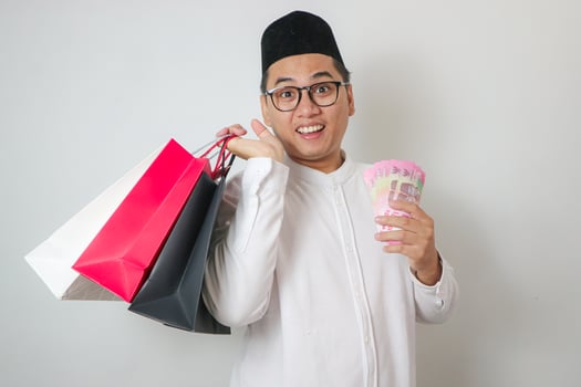 Cheerful Asian Muslim Man holding money rupiah While Shows A Few Shopping Bags To Front On white Studio Wall photo