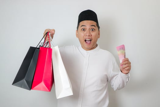 Handsome young Southeast Asian Muslim man holding rupiah money and shopping bags on isolated white studio background photo