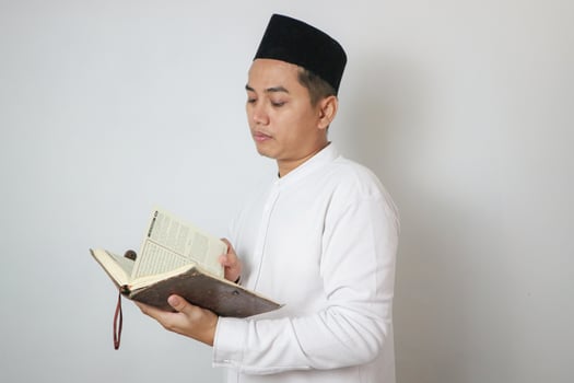 Asian Muslim man wearing white clothes reading quran during Ramadan Eid Al Fitr and eid adha days celebration isolated white background. Islamic celebration photo