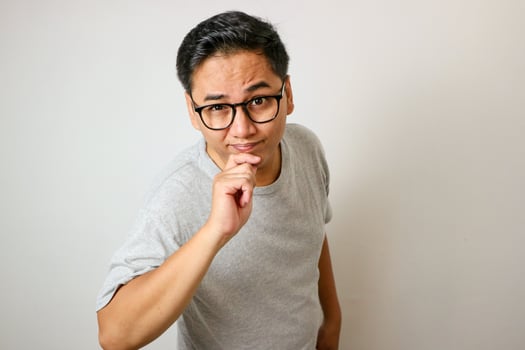 A young Asian man in a grey shirt stands thoughtfully with his hand on his chin and arms crossed, against a solid white background photo