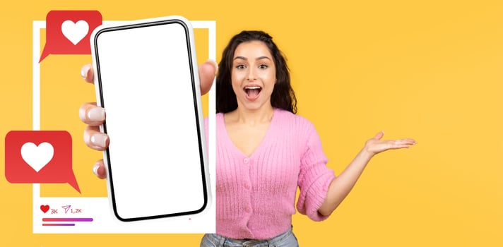 An influencer holds a smartphone displaying a blank screen, excitedly gesturing with her other hand. The vibrant yellow background enhances the lively atmosphere of online interactions. photo