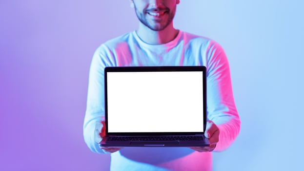 A young man holds a blank laptop in front of him, smiling brightly. The background features vibrant blue and purple lighting, creating a modern and energetic atmosphere. photo