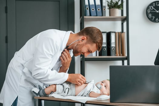 Using stethoscope. Doctor, in white coat is taking care of little toddler indoors photo