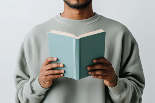 Man in a soft green sweatshirt holding a light blue book while reading in a minimalist indoor setting photo
