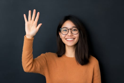 Young woman with glasses smiling and waving against a dark background in an indoor setting photo