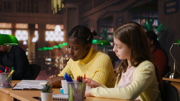 Preteen girls focus on reading required books at study desk, developing academic skills and literary language at library. Scholars enjoy lecture for school curriculum. Camera B. photo