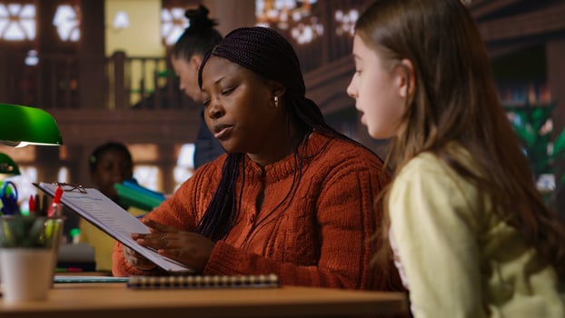 Black tutor mentoring schoolgirl in school library study desk, boosting her understanding of subject through personalized guidance. Professor hosts private lessons with pupil. Camera B. photo