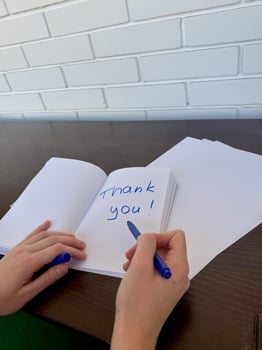 A close-up view of a young woman's right hand writing Thank You in blue marker on a notebook, against the background of a dark wooden table with white brick wall photo
