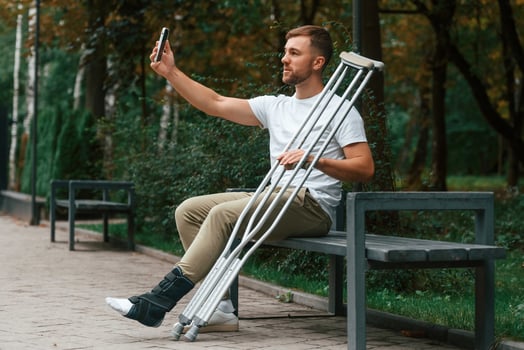 Using smartphone, to make selfie. Man with crutches is in the park outdoors. Having leg injury photo