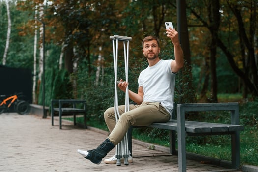 Using smartphone to make selfie. Man with crutches is in the park outdoors. Having leg injury photo