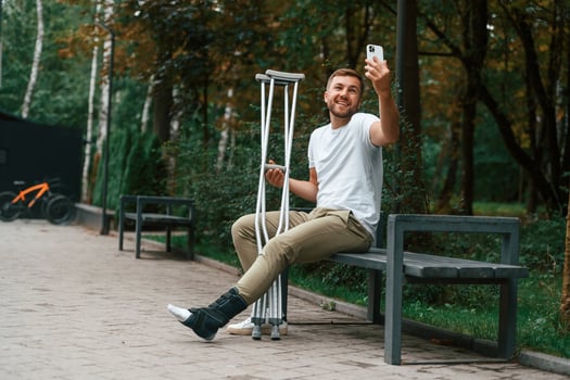 Using smartphone to make selfie. Man with ,crutches is in the park outdoors. Having leg injury photo