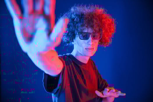 In sunglasses, showing the hand. Young man with curly hair is indoors illuminated by neon lighting photo