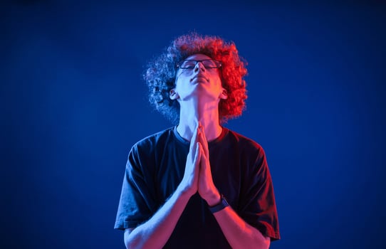 Holding hands in praying position. Young man with curly hair is indoors illuminated by neon lighting photo