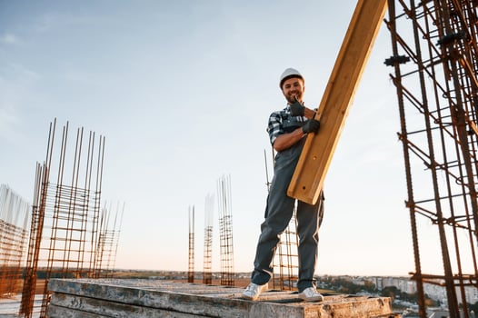 In, grey uniform. Holding wooden plank. Man is working on the construction site at daytime photo