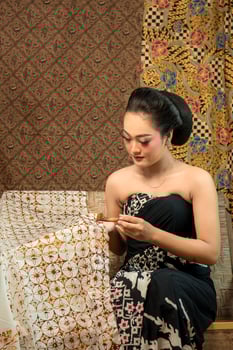 An Indonesian young woman in traditional attire making batik using a canting. She sits gracefully on a wooden bench, surrounded by handcrafted batik fabrics, showcasing cultural heritage. photo