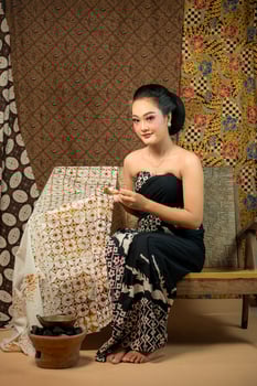 a Indonesian woman in traditional attire making batik using a canting. She sits gracefully on a wooden bench, surrounded by handcrafted batik fabrics, showcasing cultural heritage. photo