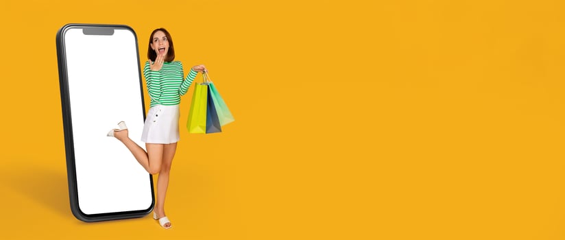 A cheerful buyer stands beside a smartphone with a big screen, excitedly holding colorful shopping bags. The background is bright yellow, emphasizing online shopping and modern commerce. photo