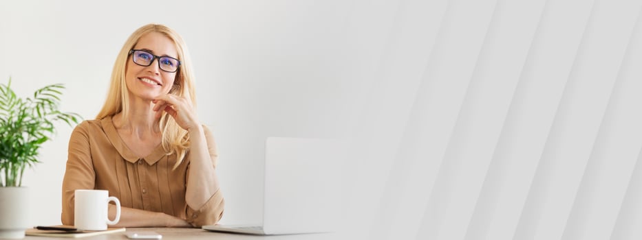 A woman with long blonde hair and glasses sits at her neatly organized desk in a home office. She is smiling while resting her chin on her hand, surrounded by a laptop and a coffee mug, copy space photo