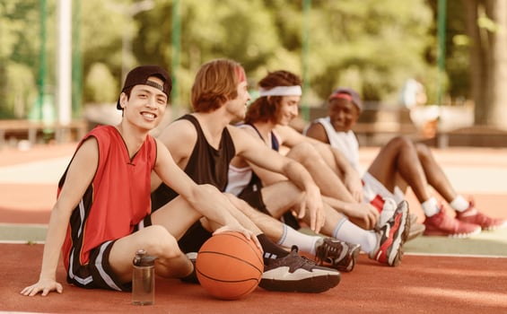 Smiling Asian basketball player with his diverse team sitting on outdoor game court, copy space. Panorama photo