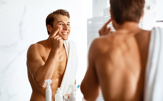 Skincare Concept. Portrait of charming shirtless guy applying cream on face, touching his cheek, looking in mirror photo