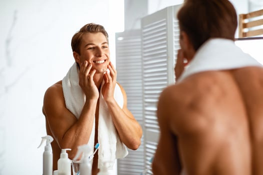 Male Facial Skincare Concept. Charming Positive Young Guy Touching His Well-Shaved Face With Smooth Skin After Shaving Routine, Shirtless Man Standing With Towel In Modern Bathroom At Home photo