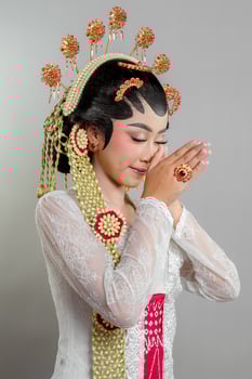 An Indonesian bride in Solo Putri attire stands gracefully with a soft smile hands in a respectful greeting. She wears a golden headdress, jasmine garland, and a white lace kebaya. photo