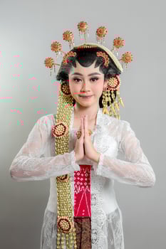 A Indonesian bride in Solo Putri attire stands gracefully with a soft smile hands in a respectful greeting. She wears a golden headdress, jasmine garland, and a white lace kebaya. photo