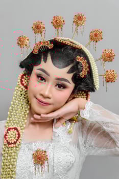 Indonesian bride in Solo Putri attire poses gracefully, resting her chin on her hand. She wears an intricate golden headdress, jasmine garland, and elegant lace kebaya photo