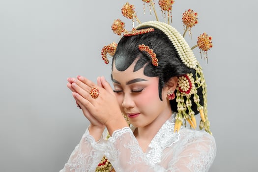 An Indonesian bride in Solo Putri attire stands gracefully with a soft smile, hands in a respectful greeting. She wears a golden headdress, jasmine garland, and a white lace kebaya photo